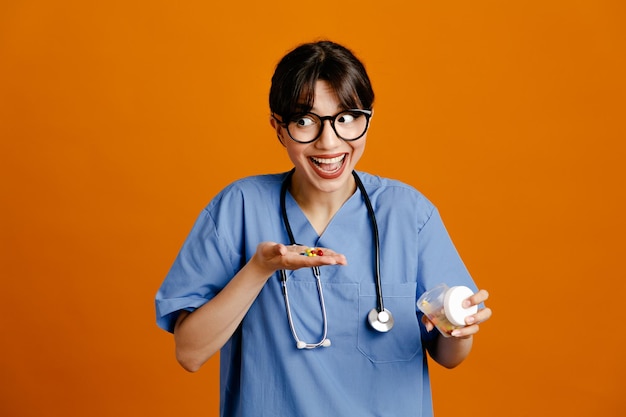 Sorridente tenendo la pillola e il contenitore della pillola giovane dottoressa che indossa uno stetoscopio fith uniforme isolato su sfondo arancione