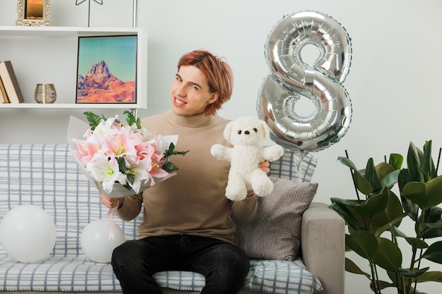 Sorridente testa inclinata bel ragazzo il giorno delle donne felici che tiene bouquet con orsacchiotto seduto sul divano in soggiorno