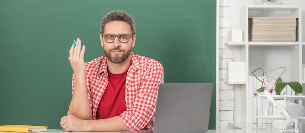 Sorridente tutor uomo con gli occhiali con laptop studente adulto studio online insegnante privato in classe con lavagna al pc torna a scuola educazione online concetto giorno della conoscenza giornata degli insegnanti
