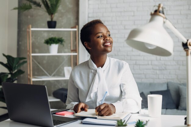 Sorridente ufficio nero donna seduta al computer portatile a prendere appunti al lavoro tecnologia internet afro americano ...