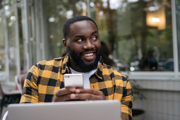 Sorridente uomo africano che tiene smartphone, lavorando in linea. Ritratto di giovane sviluppatore di successo pianificazione avvio, seduto al posto di lavoro.