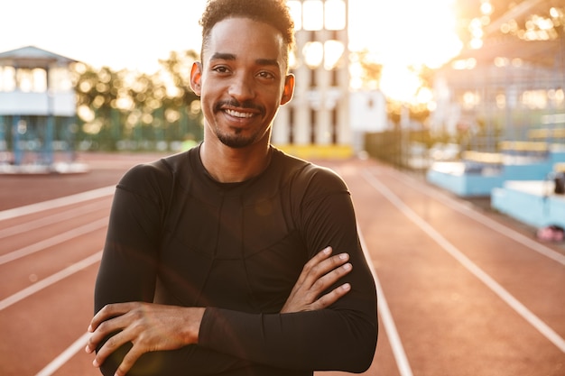 Sorridente uomo afroamericano in piedi sulla pista da corsa sul campo sportivo all'aperto