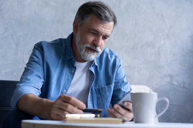 Sorridente uomo anni '50 dai capelli grigi paga le bollette online utilizzando un moderno gadget per smartphone e una carta di credito. Il cliente maturo felice della banca effettua l'acquisto di pagamento via Internet sul cellulare da casa. Concetto di tecnologia.