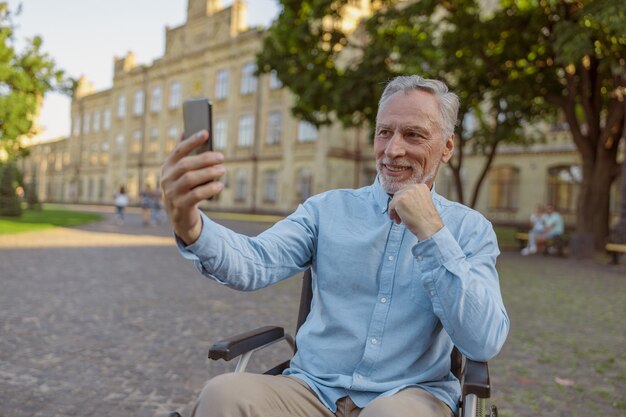 Sorridente uomo anziano che recupera un paziente in sedia a rotelle che fa una videochiamata utilizzando lo smartphone mentre