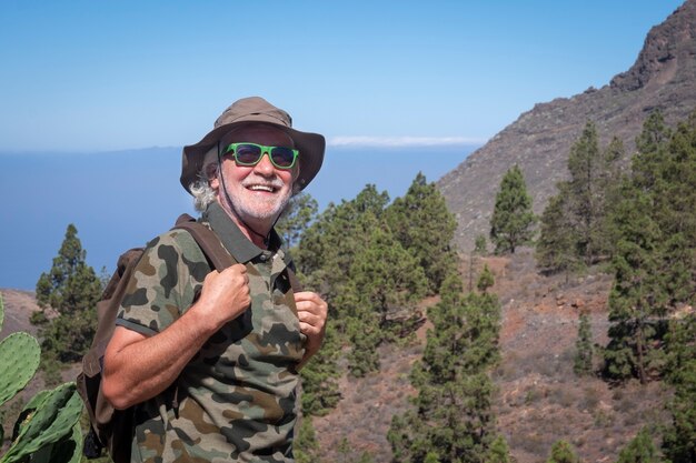 Sorridente uomo anziano con cappello e zaino che si gode un'escursione nella catena montuosa dell'isola di Tenerife - concetto di vacanza estiva per un anziano popolo attivo