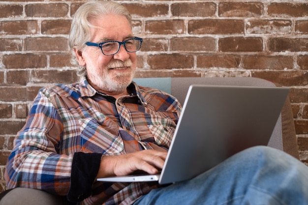 Sorridente uomo anziano rilassato seduto a casa sulla poltrona utilizzando il computer portatile Muro di mattoni