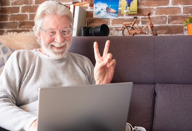 Sorridente uomo anziano utilizzando laptop a casa gesticolando segno positivo Muro di mattoni in background