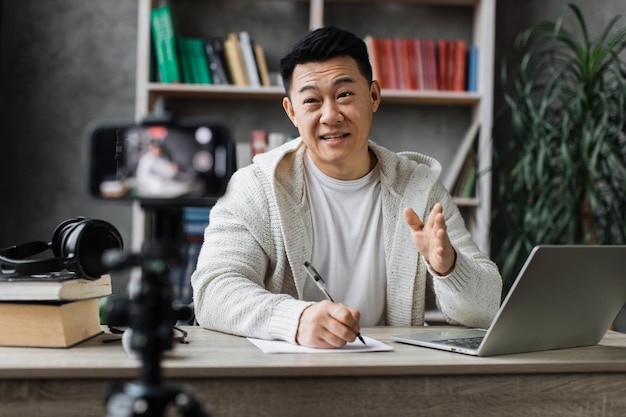 Sorridente uomo asiatico che parla e gesticola davanti al moderno rapporto di scrittura della fotocamera dello smartphone