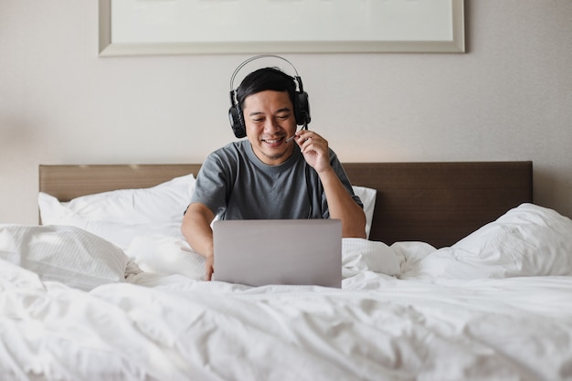 Sorridente uomo asiatico in cuffia che fa videoconferenza con il laptop sul letto