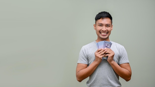 Sorridente uomo asiatico in possesso di banconote da un dollaro in contanti USD mentre in piedi contro uno sfondo verde studio