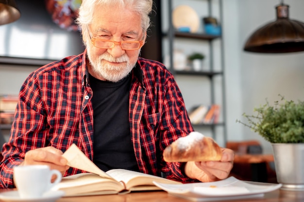 Sorridente uomo barbuto anziano seduto al tavolo del bar a leggere un libro mentre si gusta la colazione