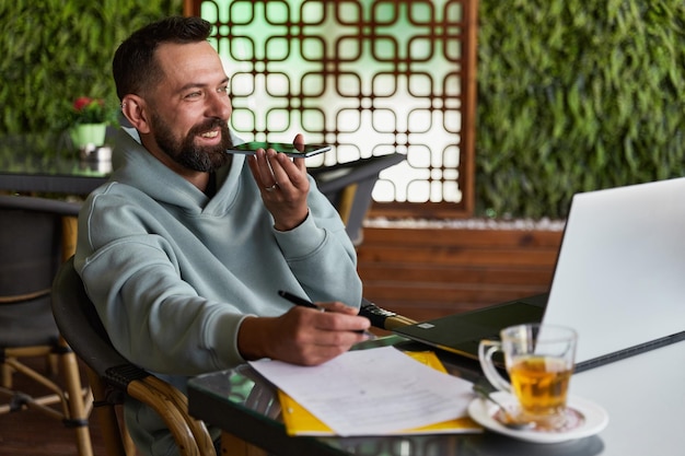 Sorridente uomo barbuto in abiti casual in caffè registra messaggi audio