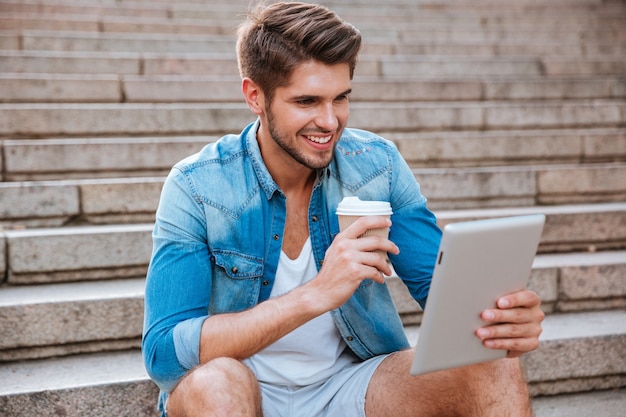 Sorridente uomo casual felice che utilizza tablet pc e beve caffè all'aperto sulla scala