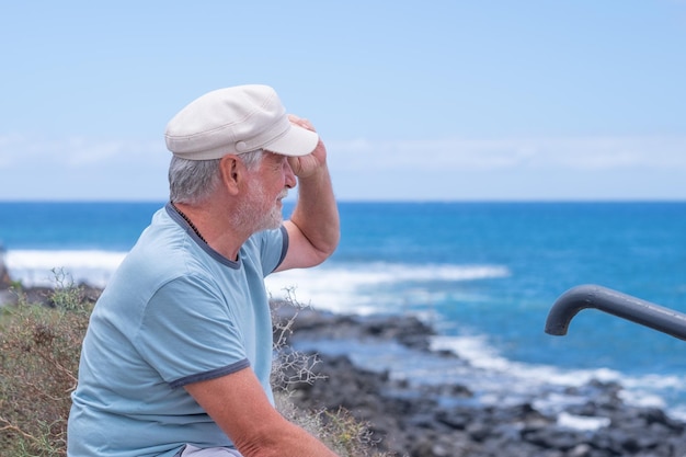 Sorridente uomo caucasico barbuto con cappello seduto in spiaggia guardando l'orizzonte mentre si gode la vacanza o il pensionamento Orizzonte sull'acqua spazio di copia