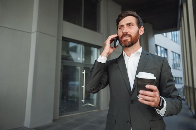 Sorridente uomo d'affari europeo in tuta con caffè che parla al telefono vicino all'edificio per uffici e distoglie lo sguardo