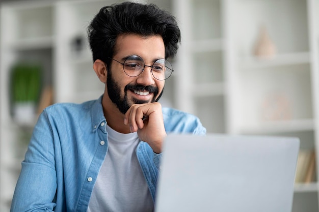 Sorridente uomo indiano in occhiali che lavora con il computer portatile in ufficio a casa