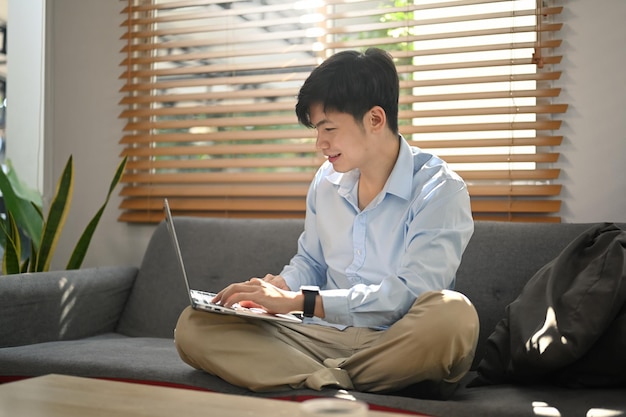 Sorridente uomo millenario seduto sul divano e che utilizza il laptop controllando la posta elettronica lavorando a distanza da casa