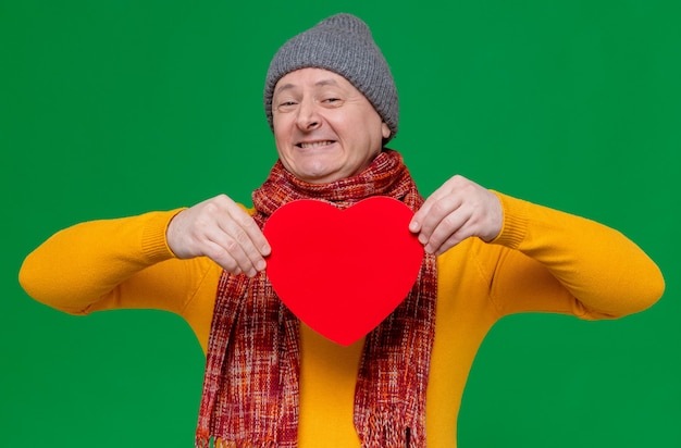 Sorridente uomo slavo adulto con cappello invernale e sciarpa intorno al collo che tiene a forma di cuore rosso e