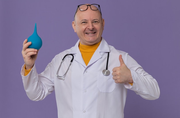 Sorridente uomo slavo adulto con occhiali ottici in uniforme da medico con stetoscopio che tiene clistere e pollice in alto