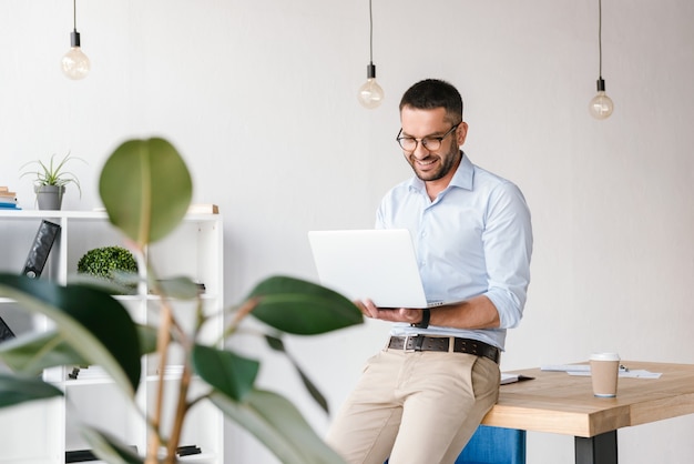 Sorridente uomo soddisfatto 30 anni indossa una camicia bianca seduto sul tavolo in ufficio e avendo chat di lavoro sul computer portatile d'argento