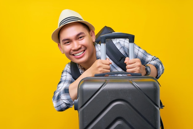 Sorridente viaggiatore turistico uomo asiatico in abiti casual cappello e zaino nascosto con valigia grigia valigia su sfondo giallo Viaggio di passeggeri all'estero nei fine settimana fuga Concetto di viaggio aereo