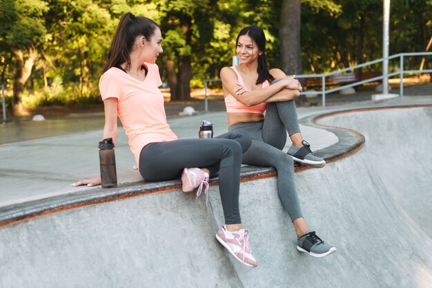 sorridenti belle donne in abiti sportivi seduti e parlando con bottiglie d'acqua sul campo sportivo in cemento