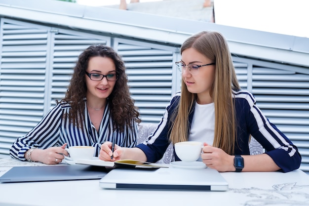 Sorridenti lavoratrici di successo stanno lavorando insieme, lavorando su un laptop sulla terrazza, colleghe sono impegnate a discutere idee, ragazze in un bar durante una riunione di lavoro. Concetto di affari