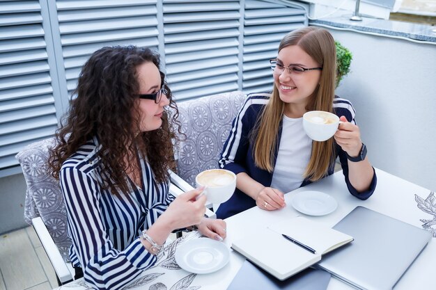 Sorridenti lavoratrici di successo stanno lavorando insieme, lavorando su un laptop sulla terrazza, colleghe sono impegnate a discutere idee, ragazze in un bar durante una riunione di lavoro. Concetto di affari