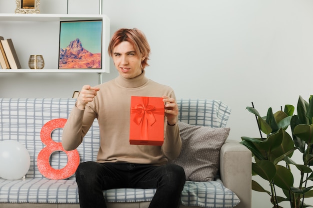 Sorridenti punti davanti a un bel ragazzo durante la giornata delle donne felici che tiene il regalo seduto sul divano in soggiorno