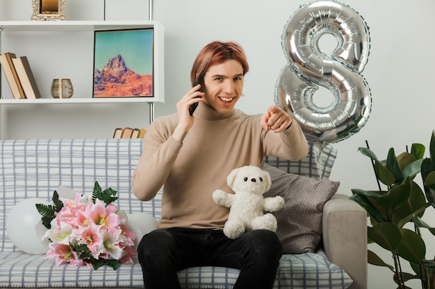 Sorridenti punti davanti a un bel ragazzo durante la giornata delle donne felici che tiene l'orsacchiotto parla al telefono seduto sul divano nel soggiorno