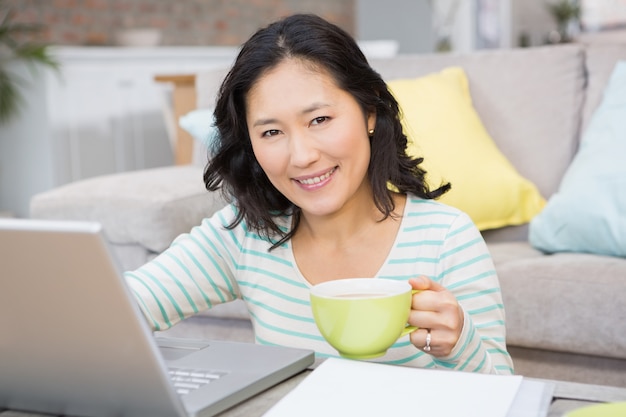 Sorridere castana facendo uso del computer portatile e della tazza di tenuta nel salone