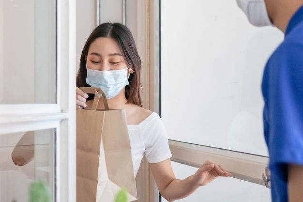 Sorridi, Felice giovane donna o ragazza asiatica che indossa una maschera per il viso protegge dalla pandemia di covid-19 e ha ricevuto un sacchetto di carta dal fattorino, messaggero in uniforme durante il blocco, quarantena alla porta di casa.