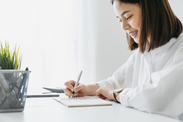 Sorridi la giovane scrittura asiatica della mano della donna sul taccuino in bianco sulla tavola in casa.