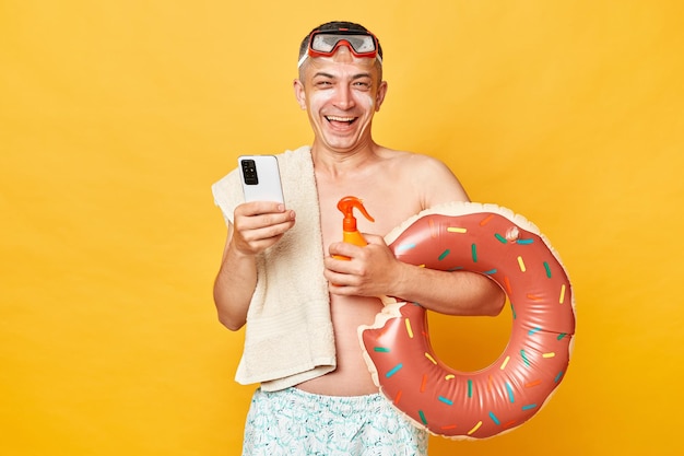 Sorrido positivo uomo turistico gioioso in località estiva in piedi con anello di gomma isolato su sfondo giallo usando il telefono cellulare mentre si rilassa vicino al mare