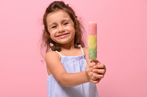 Sorrisi allegri divertenti della bambina che mostrano alla macchina fotografica un succo congelato dolce delizioso, ghiacciolo, gelato nelle sue mani. Concetto estivo per pubblicità, sfondo rosa, copia spazio per la promozione