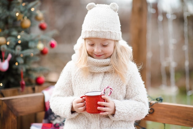 Sorriso carino kid ragazza 4-5 anni bere cioccolata calda con caramella rossa stick in tazza sopra l'albero di Natale