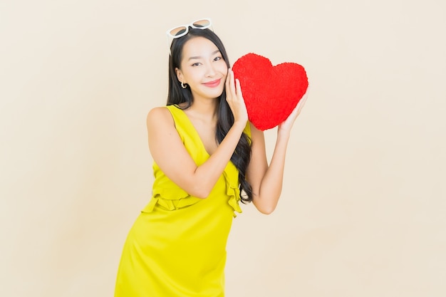 Sorriso della bella giovane donna asiatica del ritratto con la forma del cuscino del cuore sulla parete di colore