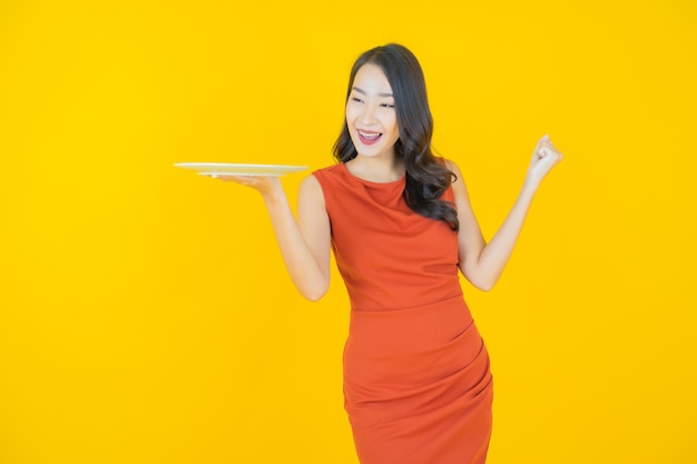 Sorriso di bella giovane donna asiatica del ritratto con il piatto vuoto del piatto su yellow