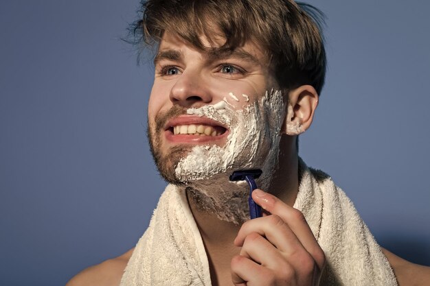 Sorriso di laurea con rasoio e asciugamano per crema da barba Uomo felice radersi il viso barbuto su sfondo blu Bellezza toelettatura igiene Cura della pelle salute benessere Concetto di routine mattutina