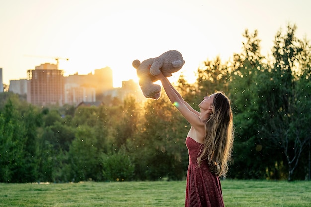 Sorriso enigmatico della donna abbronzata dai capelli lunghi carina in un vestito rosso lancia (lancia) un giocattolo di orsacchiotto grigio nelle mani sullo sfondo degli alberi del parco favolosamente atmosfera