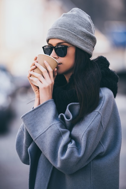 Sorso di caffè fresco. Ritratto di bella giovane donna in occhiali da sole che beve caffè e distoglie lo sguardo mentre sta in piedi all'aperto