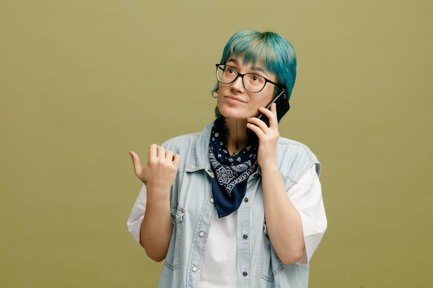 Sospettosa giovane donna che indossa occhiali bandana sul collo tenendo la mano in aria guardando in alto mentre si parla al telefono isolato su sfondo verde oliva