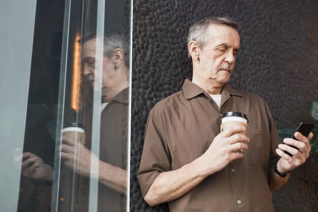 Sotto la vista di un serio manager caucasico anziano impegnato con caffè da asporto in piedi al muro nero e leggendo il messaggio sul telefono