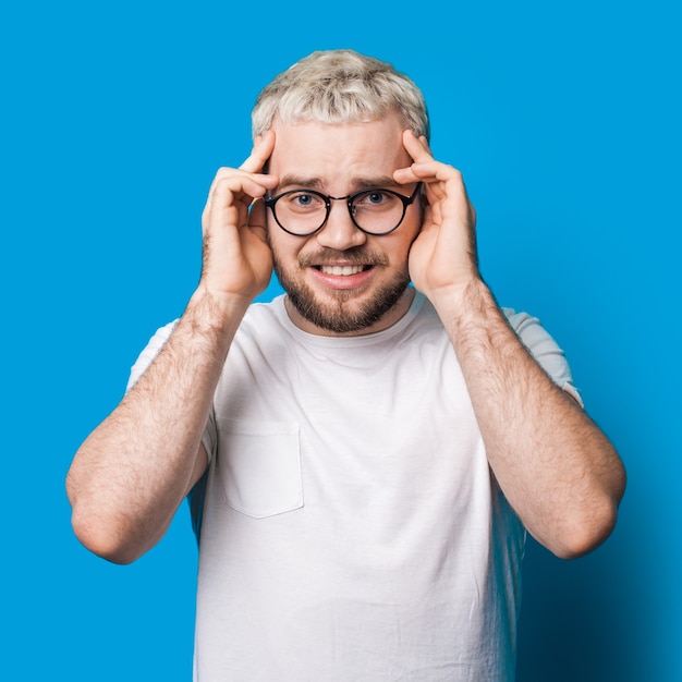 Sottolineato uomo barbuto con capelli biondi e occhiali da vista che tocca la sua testa su una parete blu dello studio