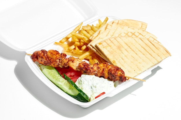 Souvlaki greco appetitoso con cetrioli, patatine fritte, pita, yogurt in una scatola di plastica per il pranzo. Cibo di strada, luce dura. Superficie bianca