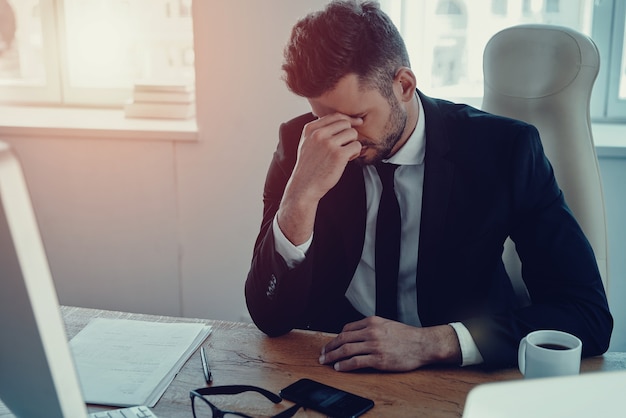 Sovraccaricato di lavoro. Stanco giovane in abiti da cerimonia che massaggia il naso mentre è seduto in ufficio