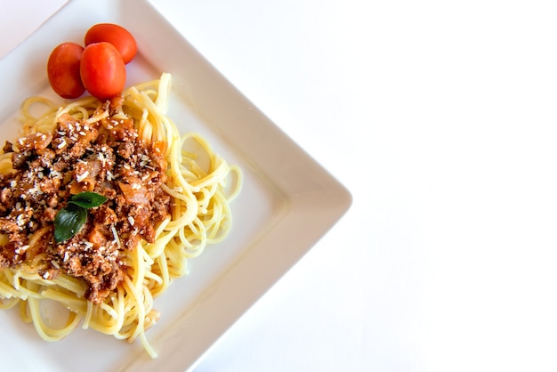 Spaghetti alla bolognese serviti su un piatto bianco su sfondo bianco con pomodori e basilico.