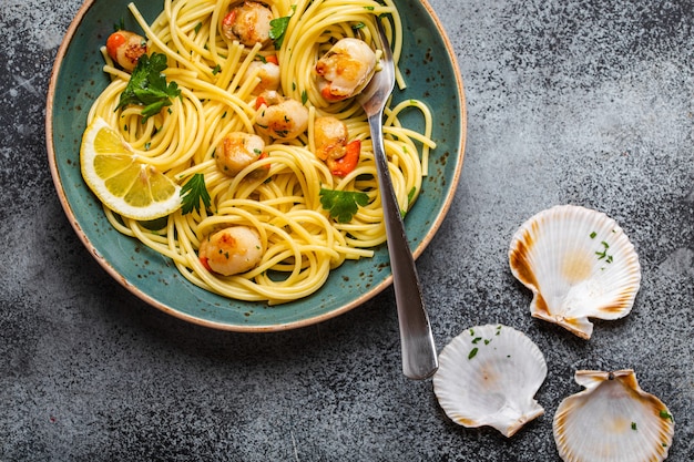 Spaghetti con frutti di mare, deliziose capesante cotte fritte servite su piatto blu, fondo di cemento rustico grigio, vista dall'alto, primo piano. Concetto di pasta ai frutti di mare