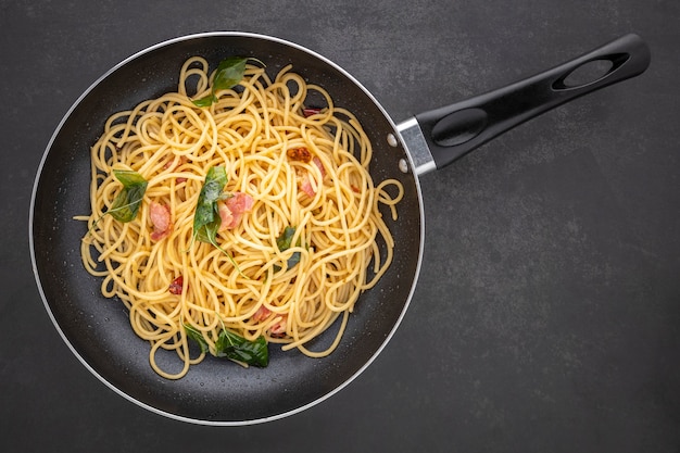 Spaghetti con peperoncino secco, aglio, basilico dolce e pancetta in padella nera, vista dall'alto