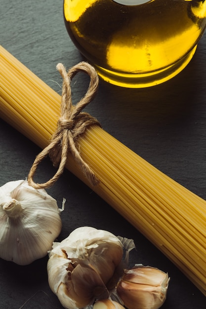 Spaghetti legati in un mazzetto adagiati sulla superficie di ardesia nera tra una bottiglia di olio e bulbi di aglio